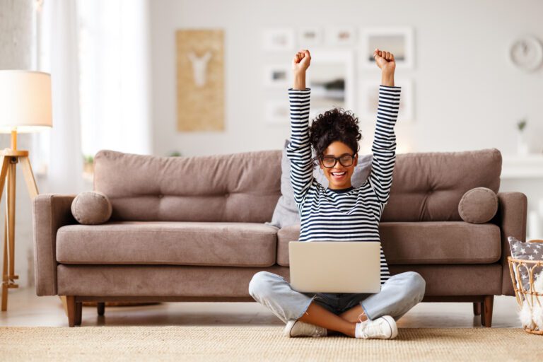 A woman sitting on the floor, raising her arms in excitement while working on her laptop.